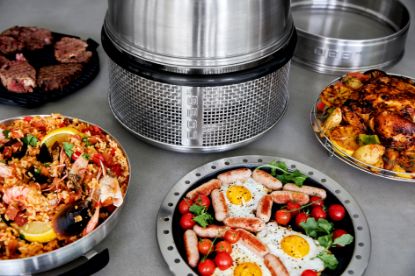 Cobb Premier Air portable charcoal cooker on a table with a roast chicken on the roasting rack, Paella in the fry dish, Sausage and eggs in the fry pan and Steaks on the griddle